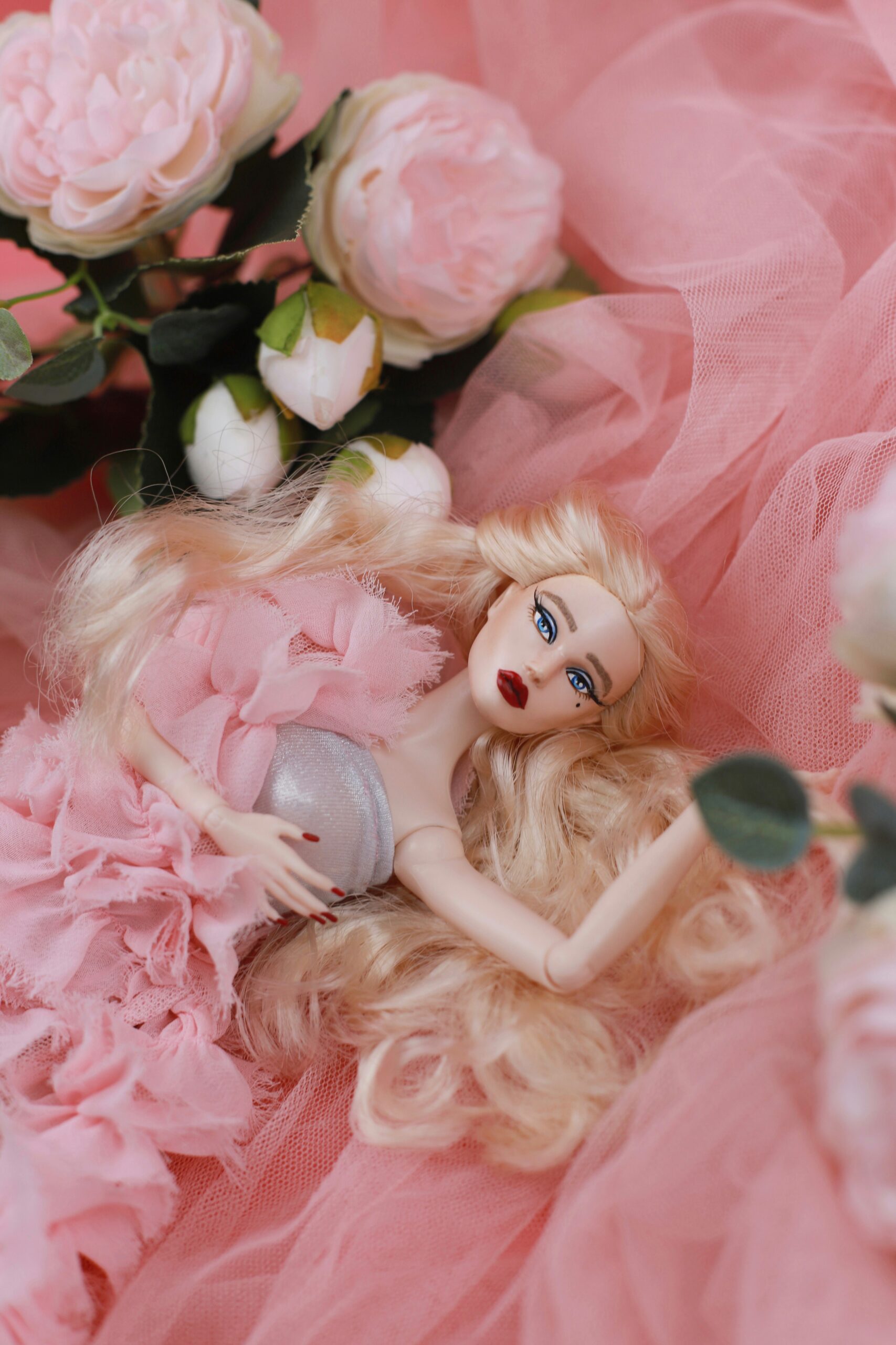 A fashion doll with long blonde hair and bright blue eyes lies on soft pink fabric, surrounded by pale pink roses. She wears a silver top with a pink ruffled gown, creating a romantic and dreamy atmosphere.